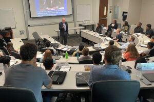 Image of Richard M. Bowen in front of the UTD MBA program with a slide of Citigroup testimony behind him during his talk in 2022.