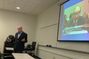 Image of Richard M Bowen speaking at the front of the room with a still of Alan Greenspan speaking into a microphone on C-SPAN 2 behind him.