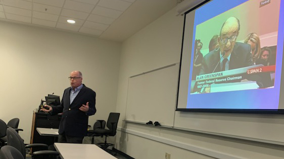 Image of Richard M Bowen speaking at the front of the room with a still of Alan Greenspan speaking into a microphone on C-SPAN 2 behind him.