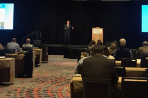 Image of Richard M. Bowen speaking on a stage before a crowd seated at tables.