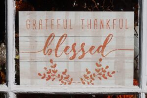 Wooden sign of white painted boards that says, "Grateful, Thankful, Blessed" in orange with orang sprigs of leaves below in a window.