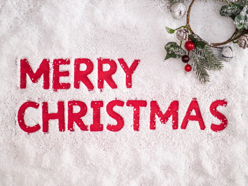 Image of the words "Merry Christmas" cut out of snow on a red surface with a holly wreath in the top right corner.