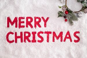 Image of the words "Merry Christmas" cut out of snow on a red surface with a holly wreath in the top right corner.