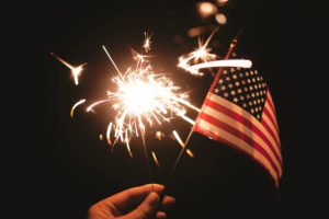 Image: hand holding sparkler and American flag with dark sky behind